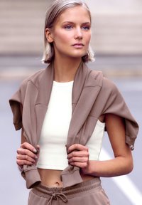 Brūna saīsināta jaka, kas valkāta virs baltas bezpiedurkņu blūzes, ar īsām piedurknēm un brīvu piegriezumu. Modeļa rokas tur jaka malas.