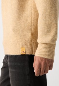 Beiger Strickpullover mit gerippten Bündchen, der eine strukturierte Oberfläche aufweist und ein gelbes Markenetikett am Saum hat. Getragen über einer dunklen Hose.