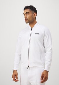 Witte sportjacket met ritssluiting aan de voorkant en zwart "BOSS" logo. Zachte stof, geribbelde manchetten en twee zijzakken. Glad textuur. Slim fit.