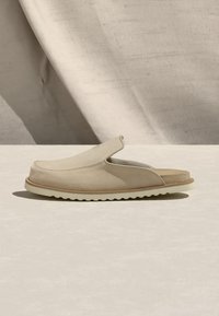 Μπεζ σανδάλι mule με μαλακό επάνω μέρος, επενδεδυμένο πάτο και υφασμάτινη λευκή σόλα, παρουσιασμένο σε ουδέτερο τόνο επιφάνειας με φόντο ύφασμα.