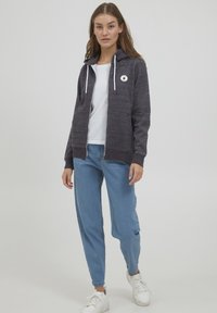 Felpa con zip di colore grigio scuro, con un logo bianco, cappuccio regolabile con cordino e tasche frontali. Abbinata a jeans azzurri chiari e sneakers bianche.