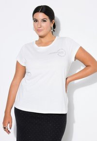 Witte t-shirt met korte mouwen en ronde halslijn met een tekstafdruk op de borst. Gecombineerd met een donkere rok met witte stippen.