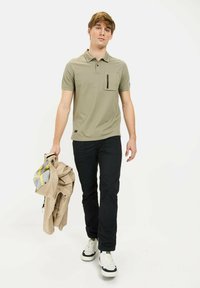 Olivgrünes Polohemd mit Brusttasche, kombiniert mit schwarzen Hosen. Das Model hält eine hellbeige Jacke. Weiße Sneaker runden das Outfit ab.