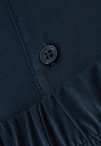 Tejido de navy con una textura suave presenta un botón negro con cuatro agujeros. La prenda incluye una sección fruncida en la parte inferior.