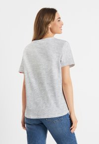 Vrouw met lang bruin haar, gekleed in een lichtgrijze korte mouwen shirt en blauwe jeans, staand en glimlachend met haar lichaam zijwaarts gericht tegen een witte achtergrond.