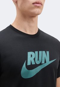 Κοντινό πλάνο ενός ατόμου που φοράει ένα μαύρο μπλουζάκι με τυρκουάζ κείμενο "RUN" και λογότυπο Swoosh στο στήθος, περικομμένο στον λαιμό και τους ώμους.