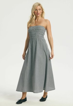 Blondína v čierno-bielych maxi šatách bez ramienok s kockovaným vzorom a čiernych balerínach stojaca pred jednoduchým pozadím.