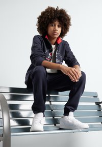 Tuta blu navy con accenti rossi, con logo sul petto. Sneakers bianche, seduto su una panchina blu metallica.