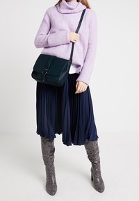 Marinblå plisserad kjol, ljuslila stickad polotröja, grå stövlar med ormskinnsmönster, och en mörkblå axelväska med rem och spänndetalj.