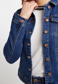 Jeansjacka i mörkblått med en åtsittande design, kopparknappar och slitna detaljer. Bärs över en ljusgrå topp med svarta ränder.