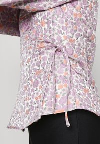 Blusa com laço floral apresentando um padrão em lavanda, laranja e cinza. Feita de tecido leve com detalhes texturizados e fendas laterais.