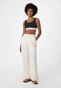 Musta crop-toppiyläosa, jossa valkoinen raidallinen nauha, yhdistetty beigeihin leveälahkeisiin housuihin, joissa on sivuhaarat ja logo. Malli seisoo neutraalin taustan edessä.