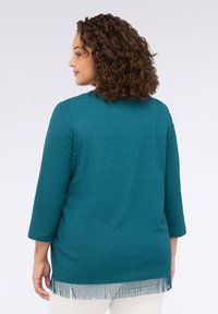 Top tricotat de culoare teal, cu mâneci trei sferturi, model texturat și detaliu din franjuri la margine, având o croială lejeră.