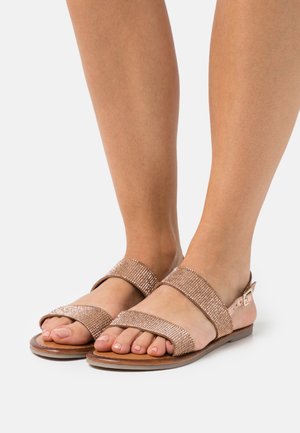 Pies con sandalias de punta abierta en oro rosa, con dos tiras brillantes y una hebilla sobre un fondo neutro.