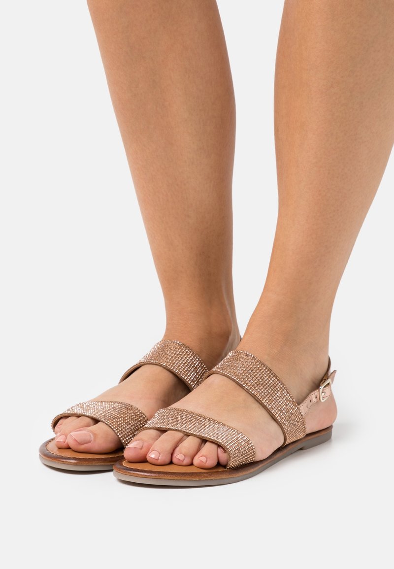 Pieds portant des sandales ouvertes rose doré avec deux brides scintillantes et une boucle sur un fond neutre.