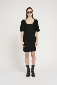 Črna obleka z oprijetim krojem in kvadratnim izrezom ter kratkimi nabranimi rokavi, v kombinaciji s črnimi gležnarji. Model nosi tudi elegantne črne sončne očala.