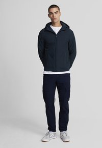 Giacca zip-up blu navy con cappuccio, abbinata a pantaloni cargo scuri. La giacca ha una texture liscia e dettagli minimi. Logo distintivo sul petto.