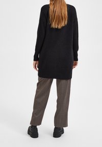 Svart, lång tunika-sweater med mjuk textur och avslappnad passform, matchad med bruna randiga byxor och svarta klumpiga stövlar.