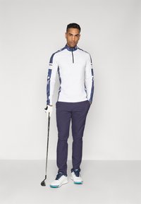 Man in navy pants and white long-sleeve golf shirt holding a golf club, standing against plain light background.