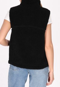 Gilet en polaire noire avec un col montant, une surface texturée et des fentes latérales. L'ourlet est arrondi, porté sur une chemise blanche et un jean bleu.