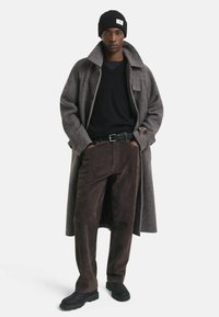 Lungo cappotto a quadri marrone-grigio, maglione nero, pantaloni in scamosciato marrone, scarpe nere, berretto nero. Sfondo semplice, enfasi sulle texture dell'abbigliamento esterno.