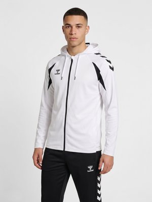Młody mężczyzna stoi, ubrany w białą kurtkę z zamkiem i kapturem oraz czarne spodnie z dopasowanym czarno-białym sportowym wzorem w kształcie litery V.