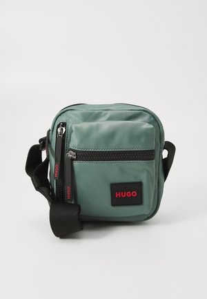 Geantă de umăr din nailon verde cu accente negre, dotată cu două compartimente cu fermoar, curea reglabilă și un patch proeminent cu logo-ul HUGO.