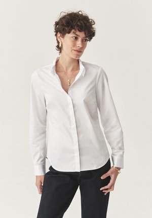 Woman with short curly hair wearing a white button-up shirt, dark pants, and gold jewelry, standing against a plain light background.