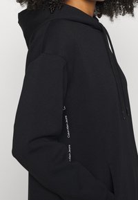 Henkilö, jolla on päällään musta Calvin Klein Jeans -huppari, jossa bränditeksti näkyy sivussa. Henkilön käsi on osittain etutaskussa.