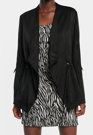 Frau, die eine schwarze Jacke mit offenem Vorderteil über einem schwarz-weißen Zebramusterkleid trägt, steht vor einem schlichten hellen Hintergrund.