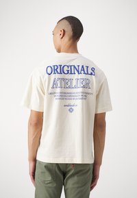 Cream short-sleeve t-shirt with blue text “ORIGINALS ATELIER” and additional branding on the back; lightweight cotton fabric, relaxed fit.