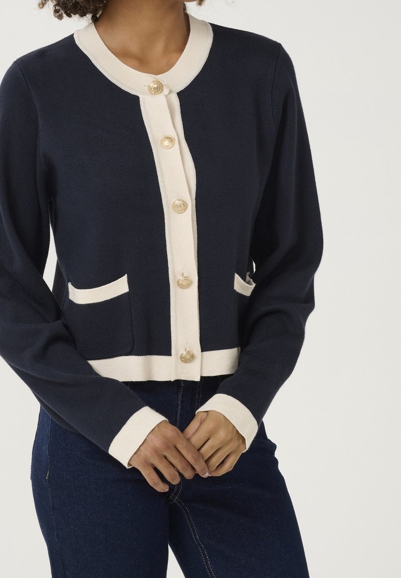 Marineblaue Strickjacke mit cremefarbener Borte und Knöpfen, ausgestattet mit zwei vorderen Taschen und einem runden Halsausschnitt. Das Design ist tailliert und hat lange Ärmel.
