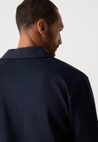 Man with short hair and beard facing sideways, wearing a dark navy blue collared shirt against a plain light background.