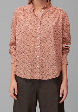 Vrouw draagt een lange mouwen, rode bloemenprint blouse met knopen en een kraag, gecombineerd met een donkerbruine broek tegen een effen achtergrond.
