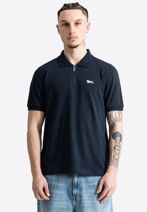 Man met gevlochten haar die een zwart polo met korte mouwen draagt met een tijgerlogo en lichtblauwe spijkerbroek, waarbij tatoeages op zijn linkerarm zichtbaar zijn.