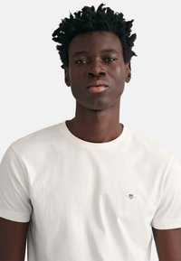 Young man with short curly hair wearing a plain white crew-neck t-shirt with a small shield logo on the chest.