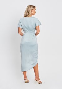 Svetlo modra satenasta obleka s kapicami, prilegajoča silhueta in diskreten visok-nizak rob. Zapiranje z zadrgo na hrbtu in gladka tekstura.