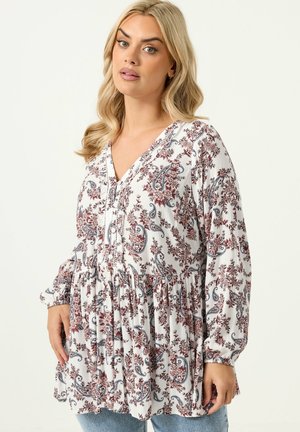 Donna con capelli biondi che indossa una tunica bianca a maniche lunghe con motivi floreali paisley rossi e blu e jeans blu.