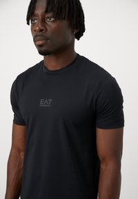 T-shirt de sport à manches courtes noir, fabriqué en tissu lisse et extensible. Présente un petit logo EA7 réfléchissant gris sur la poitrine.