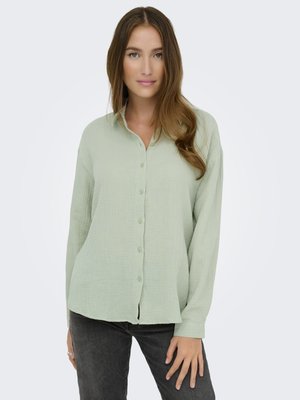 Chemise boutonnière à manches longues de couleur vert clair, en tissu texturé, offrant une coupe décontractée et un motif doux et subtil ; associée à un jean foncé.