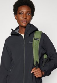 Veste zippée noire avec une capuche et des poches avant, arborant un logo sur la poitrine. Une bandoulière verte est partiellement visible.