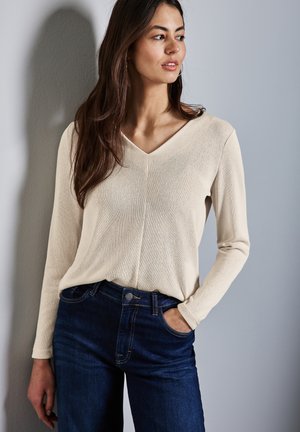 Beige langærmet top med V-udskæring og tekstureret stof, båret med mørkeblå jeans. Modellens hånd er i lommen på jeansene.
