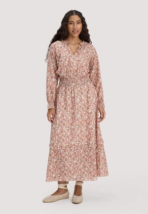 Frau mit langen lockigen Haaren, die ein langärmliges, knöchellanges Kleid mit Blumenmuster und beige geschnürte flache Schuhe trägt, vor einem schlichten Hintergrund stehend.