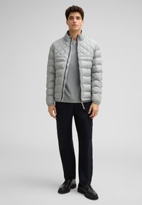 Junger Mann steht da, trägt eine hellgraue gesteppte Jacke, ein graues Hemd, schwarze Cargo-Hosen und schwarze Schnürstiefel vor einem schlichten Hintergrund.