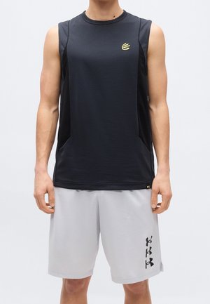 Homme portant un haut de sport noir sans manches et un short gris clair avec des logos noirs, debout devant un fond clair uni.