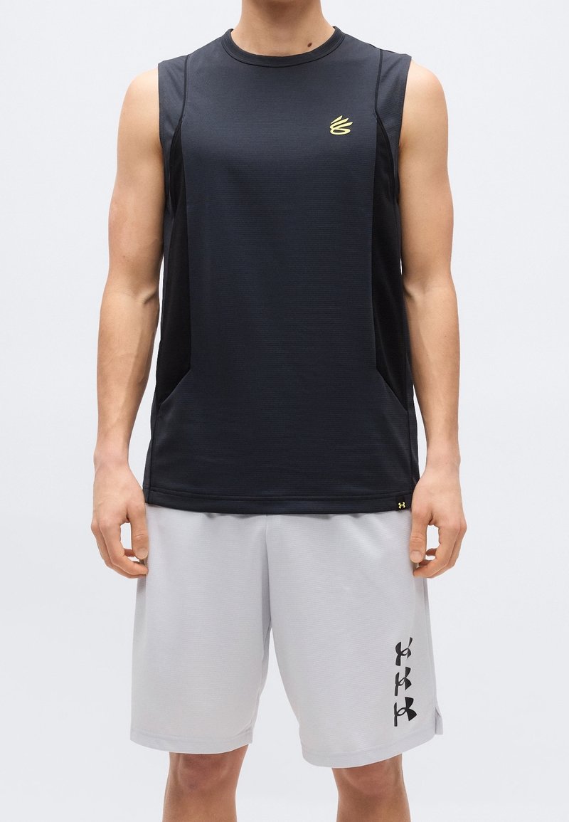 Homme portant un haut de sport noir sans manches et un short gris clair avec des logos noirs, debout devant un fond clair uni.