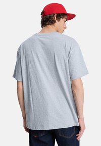 Γκρι βαμβακερό t-shirt με κοντά μανίκια, άνετη εφαρμογή και στρογγυλή λαιμόκοψη, φορεμένο με μπλε τζιν, από την πίσω όψη. Είναι ορατό επίσης ένα κόκκινο καπέλο.