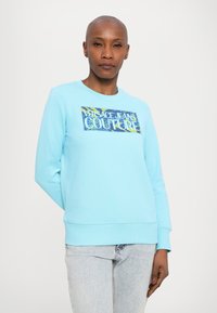 Világoskék pulóver, puha anyagból készült, kerek nyakkivágással, az elején egy színes nyomtatott logóval, melyen a "VERSACE JEANS COUTURE" felirat olvasható.