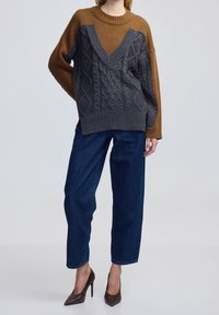 Ruskean ja harmaan sävyinen V-kaula-aukkoinen neulepusero, jossa on palmikkoneulekuvio. Yhdistetty väljiin tummiin farkkuihin ja ruskeisiin korkokenkiin.