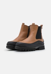 Bottines en cuir marron avec un côté élastique noir texturé, semelle en caoutchouc et lanière à l'arrière. Elles présentent une forme moderne et épurée avec des matériaux contrastés.